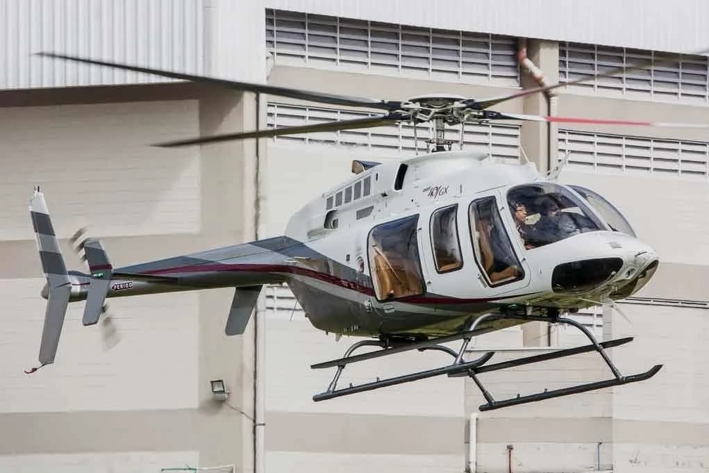 A white and grey Bell 407GX helicopter with a red stripe hovers in front of a light-colored building.
