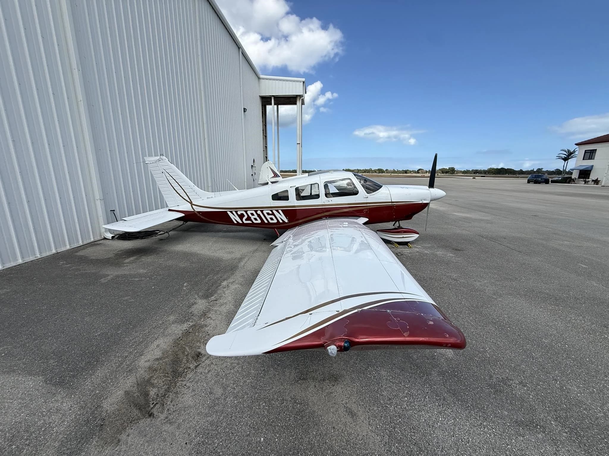 A white and maroon single-engine propeller aircraft, tail number N2816N, is parked on an asphalt apron next to a white hangar under a blue sky.