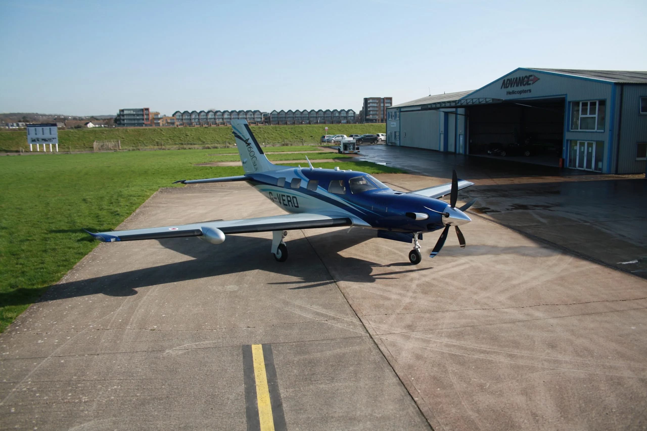 A blue and silver Piper M600SLS aircraft (G-VERO) parked on a tarmac next to an "Advance Helicopters" hangar.
