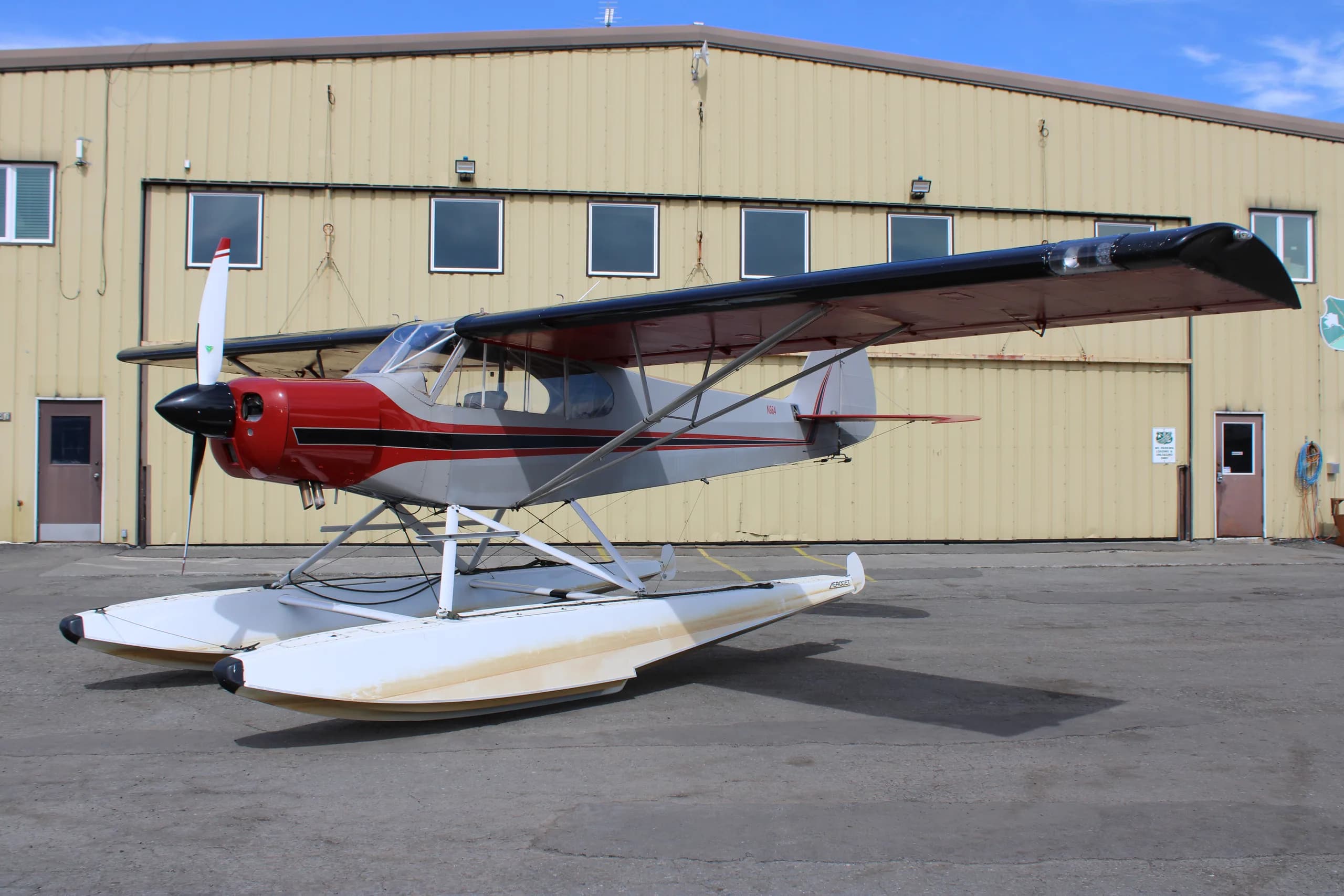 A red and silver float plane is parked in front of a hangar.