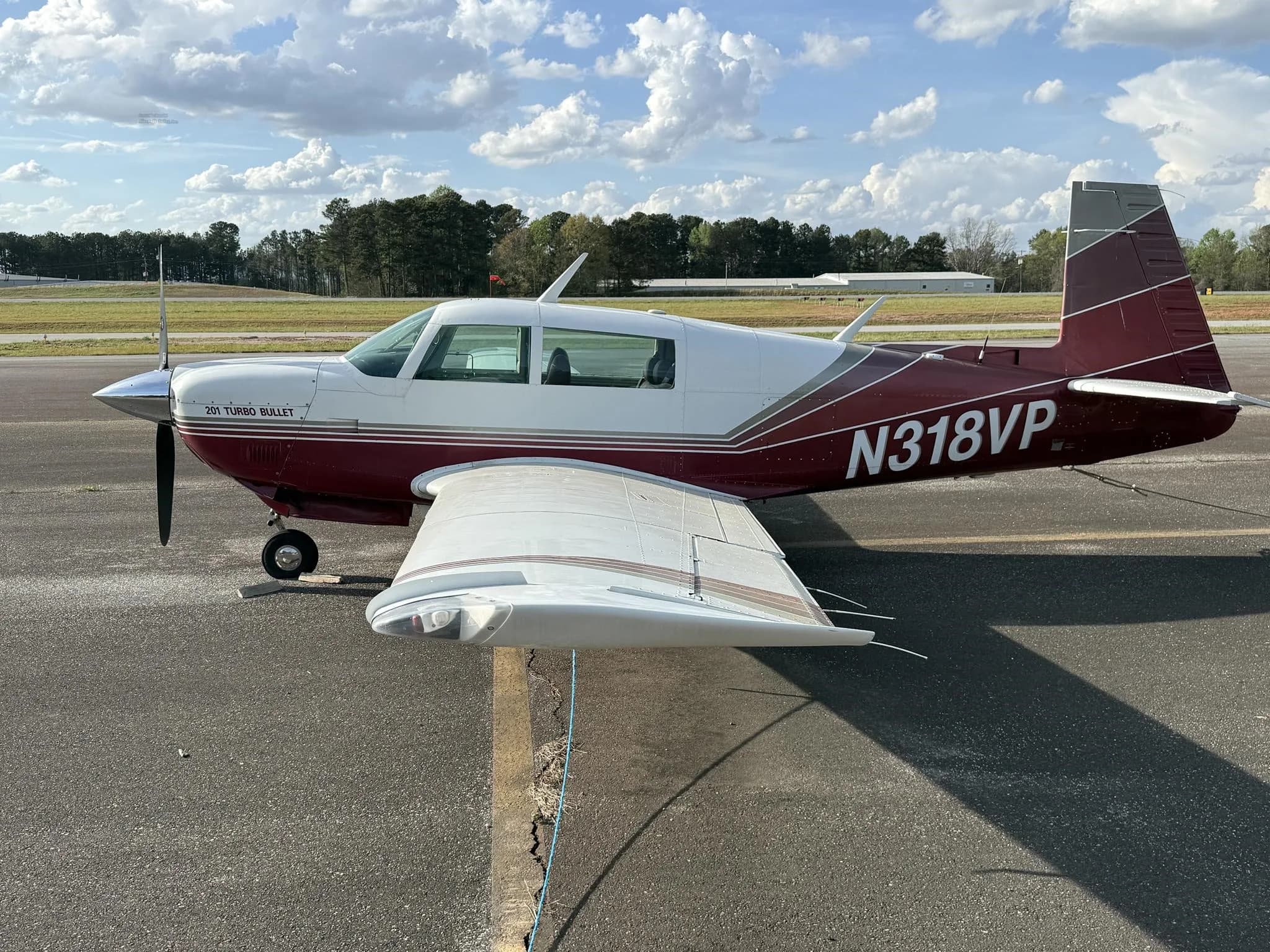 A white and maroon Mooney 201 Turbo Bullet aircraft (N318VP) is parked on an asphalt tarmac under a partly cloudy sky.