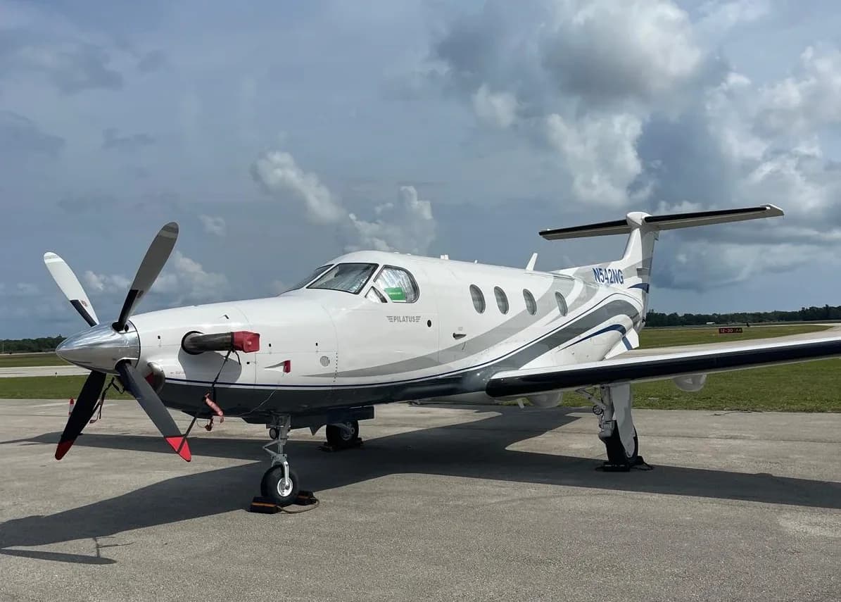 A white Pilatus PC-12 NG aircraft with blue and grey stripes, registration N542NG, parked on a tarmac under a cloudy sky.