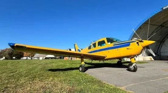 A yellow and blue single-engine aircraft parked on grass next to an asphalt path and a hangar under a clear blue sky.