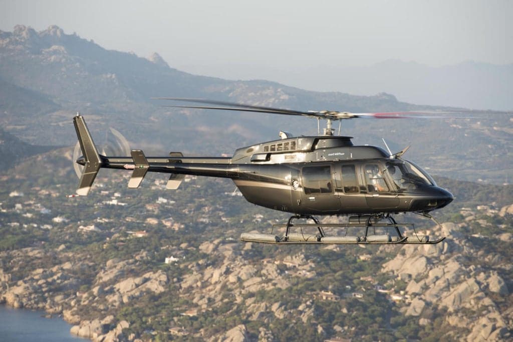 A dark grey Bell 407GXi helicopter in flight over a rocky landscape.