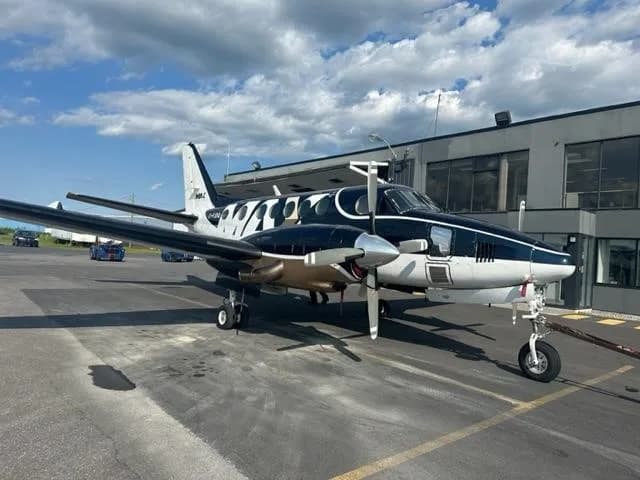 A twin-engine turboprop aircraft with a dark blue, white, and gold paint scheme, parked on an airport apron next to a building.