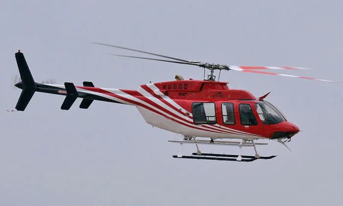 A red and white Bell 206 helicopter with a striped tail rotor in flight against a grey sky.