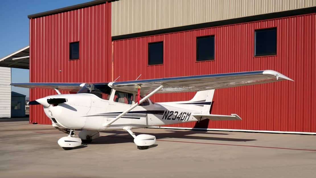 A Cessna 172 Skyhawk, tail number N234GM, is parked in front of a red hangar.