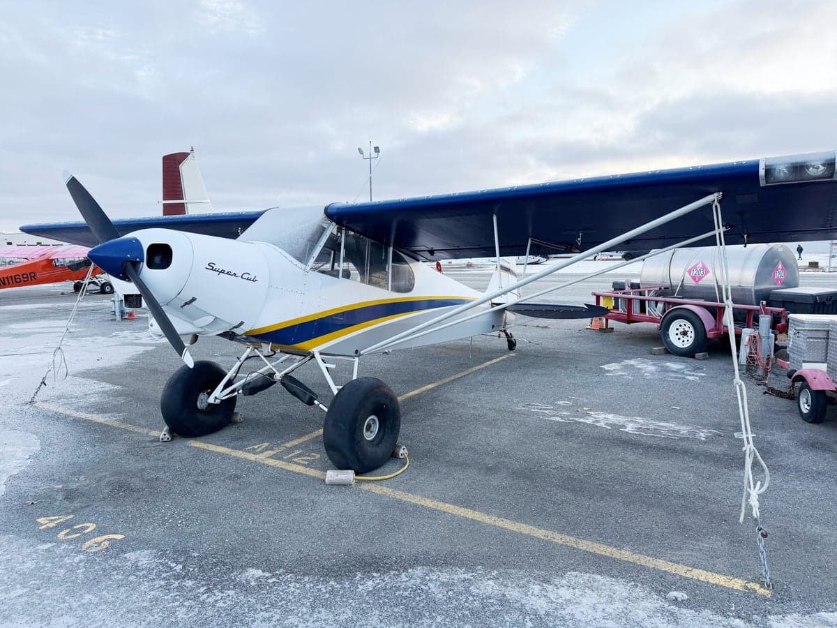A white Super Cub aircraft with blue and yellow stripes and large tundra tires, parked on a snowy tarmac next to a fuel trailer.