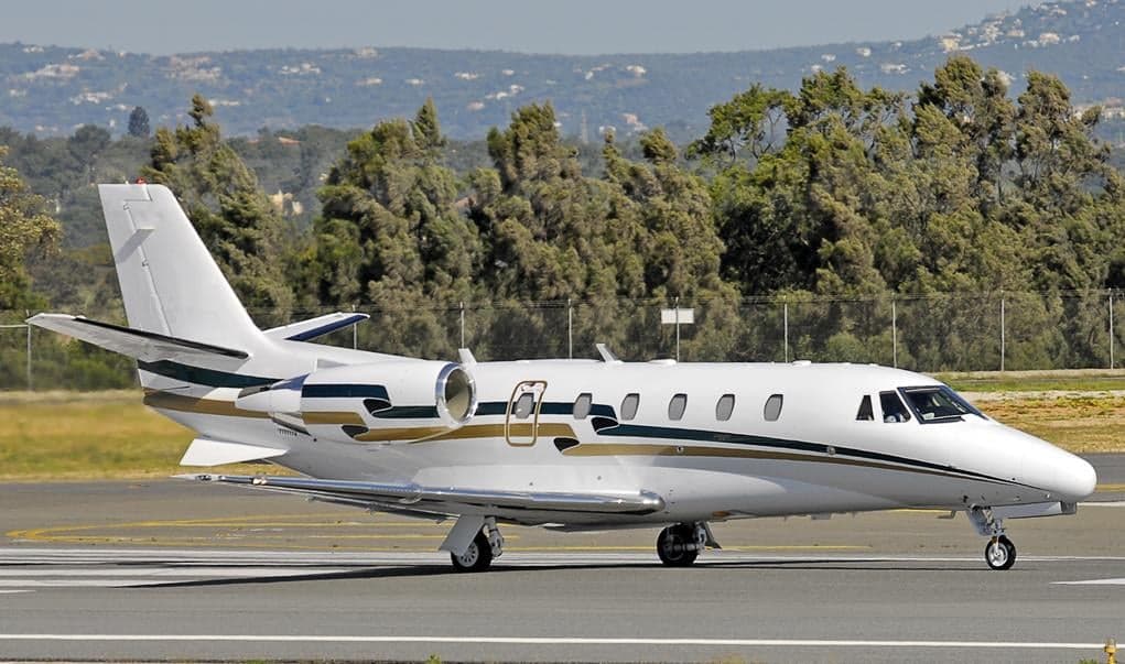 A white business jet with green and gold stripes on the fuselage is parked on an airport tarmac with trees and hills in the background.