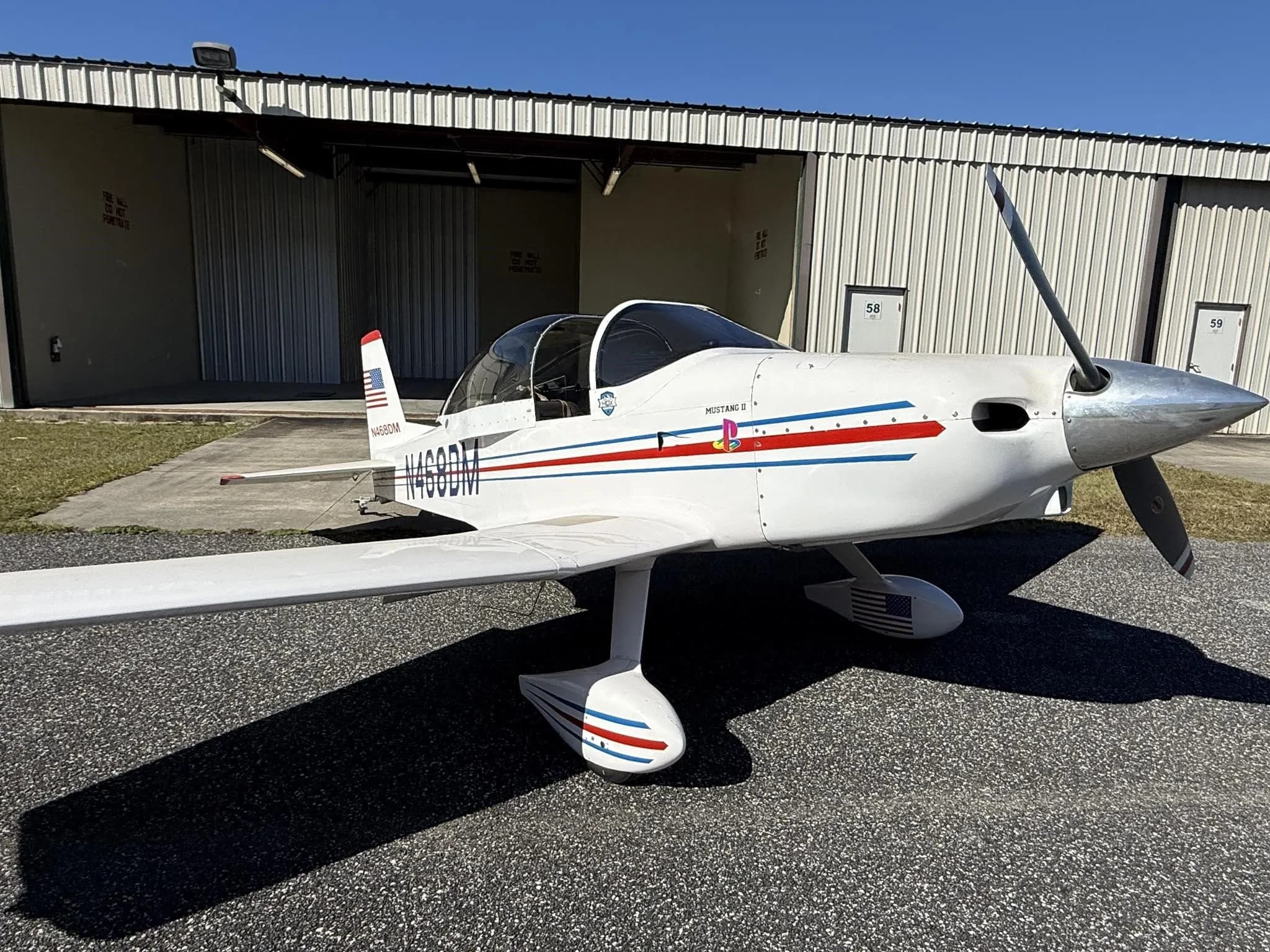 A white Mustang II aircraft with red and blue stripes, registration N468DM, parked on a tarmac in front of hangars.