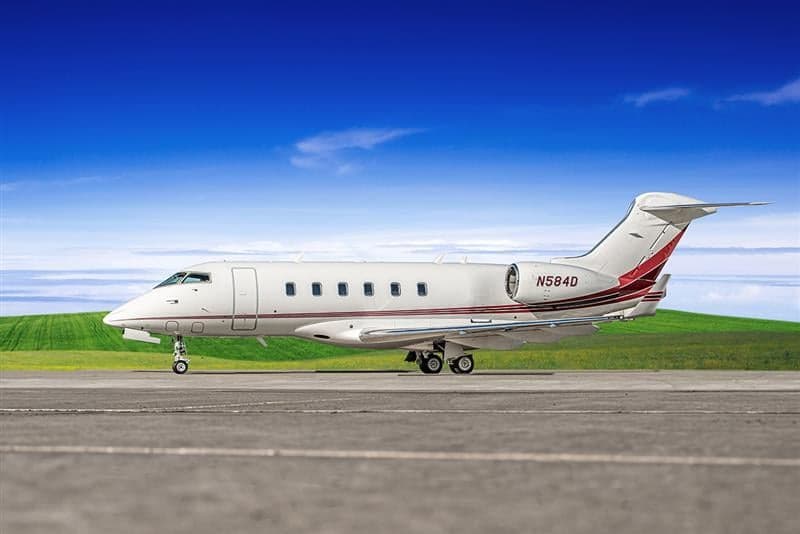 A white Bombardier Challenger business jet with red and maroon stripes, parked on an airfield.