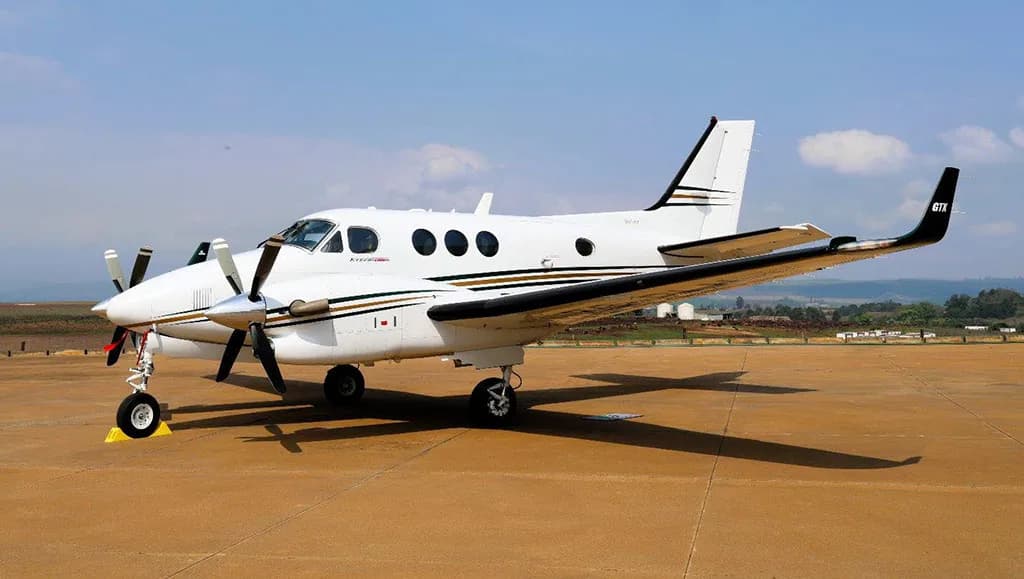 A white Beechcraft King Air C90GTx twin-turboprop aircraft with gold and black stripes parked on a tarmac under a partly cloudy sky.