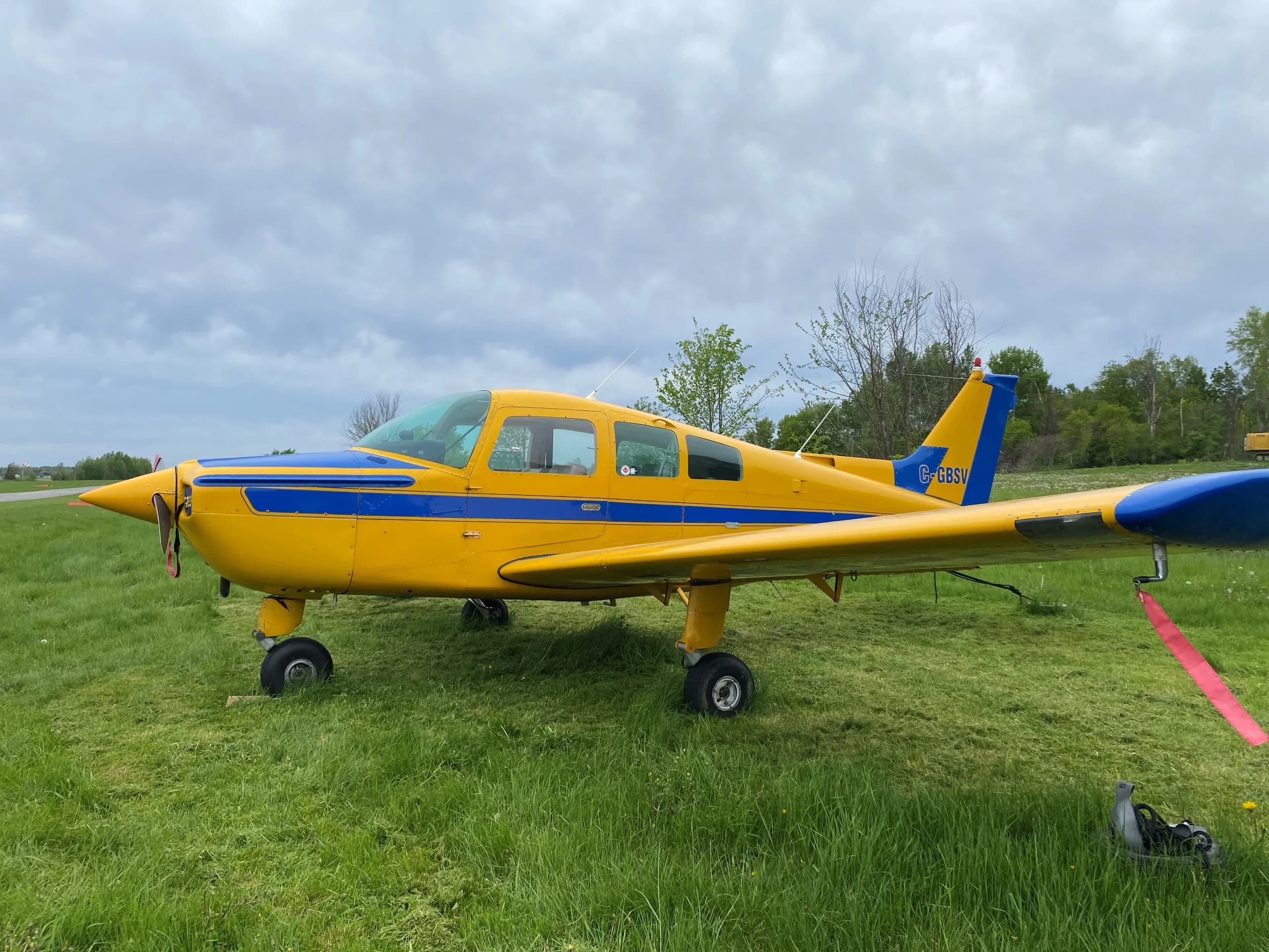 A yellow single-engine propeller aircraft with a blue stripe and tail (C-GBSV) parked on a grassy field under a cloudy sky.