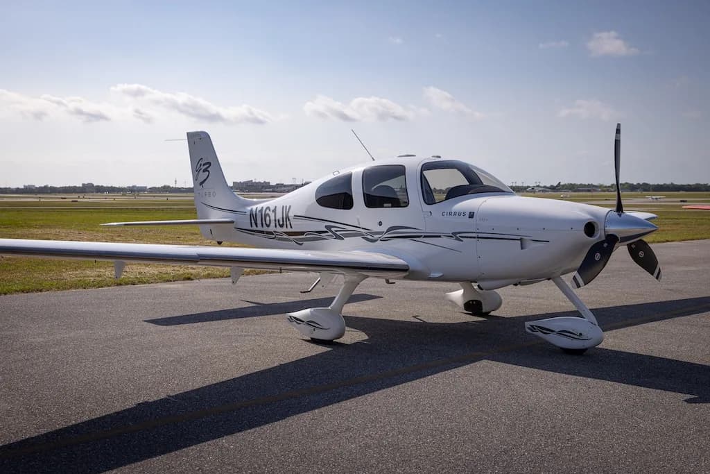 A white Cirrus SR22 Turbo aircraft with black and grey stripes, registration N161JK, parked on an asphalt tarmac under a blue sky.