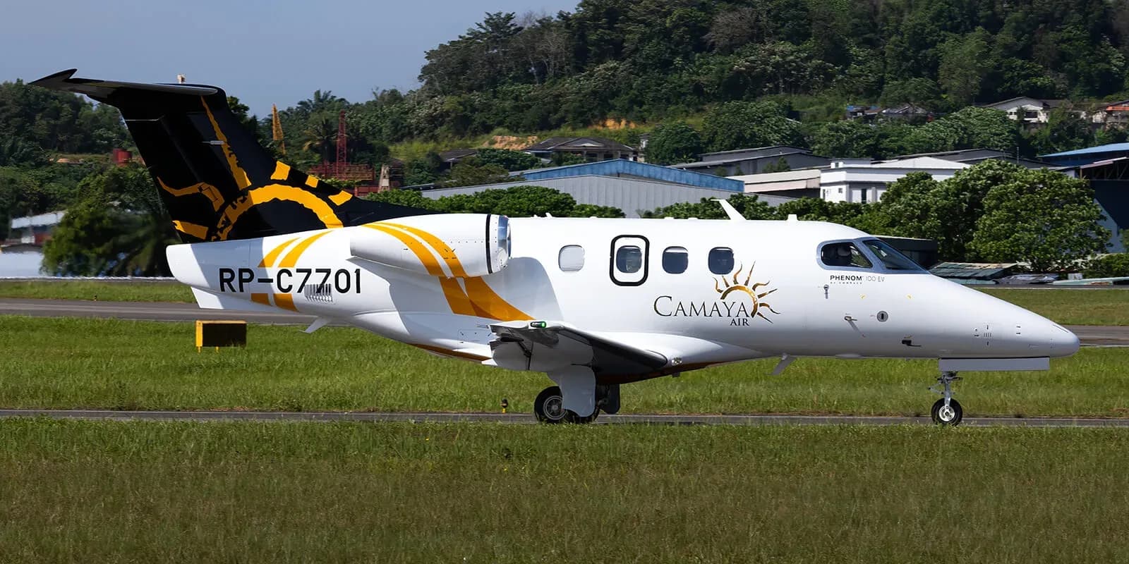 A Camaya Air Embraer Phenom 300, registration RP-C7701, on a taxiway with a jungle background.