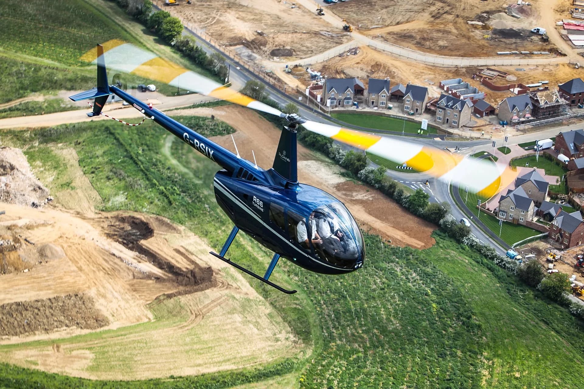 A blue Robinson R66 Turbine helicopter in flight over a suburban area.