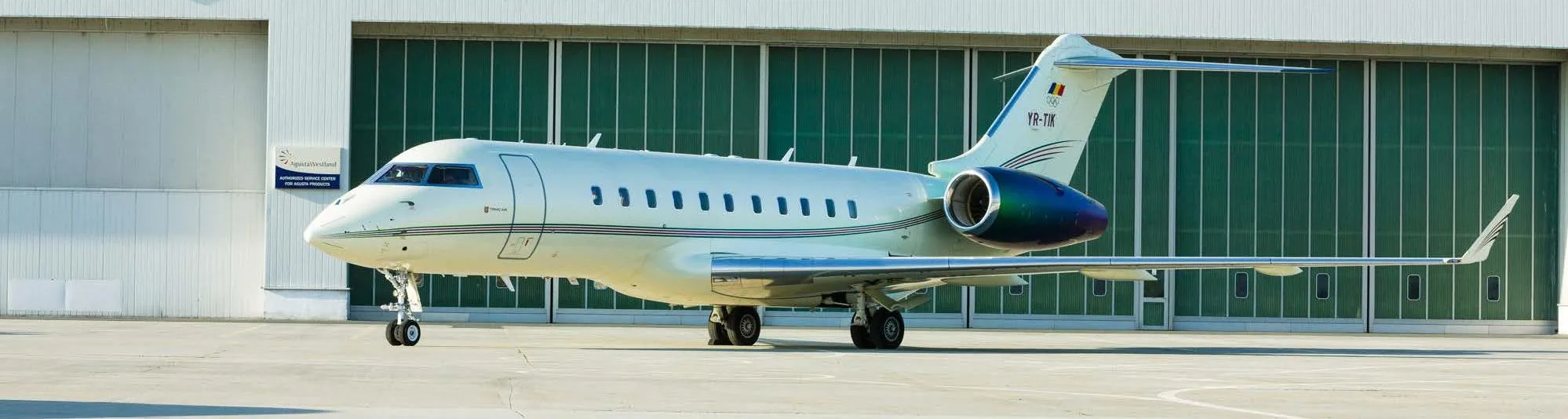 A white Bombardier Global 5000 business jet with blue and red accents is parked on a tarmac in front of a hangar.