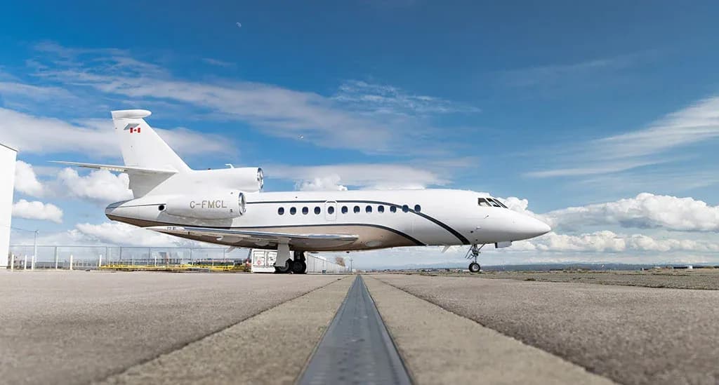 A white Dassault Falcon business jet with a brown stripe, registration C-FMCL, parked on an airfield under a blue sky.