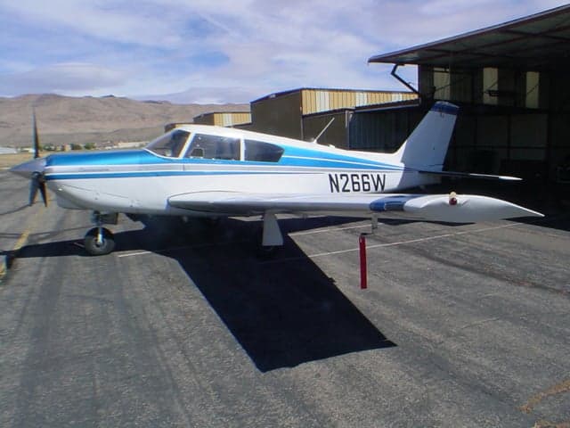 A white and blue single-engine propeller aircraft (N266W) is parked on an asphalt ramp next to hangars with mountains in the background.