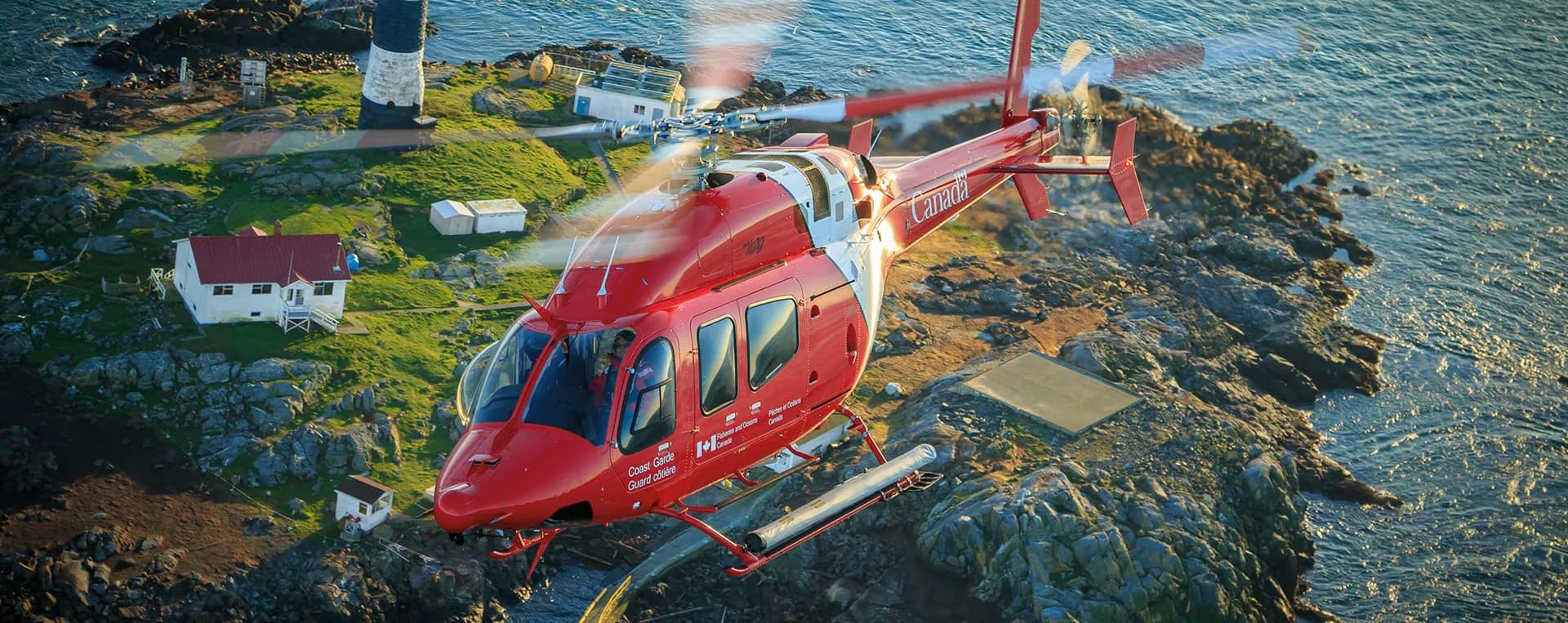 A red Bell 429 helicopter flying over a rocky coastline.