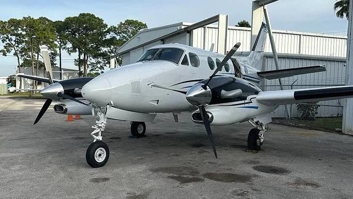 A white and black Beechcraft King Air with blue accent stripes parked on an asphalt apron next to a hangar.