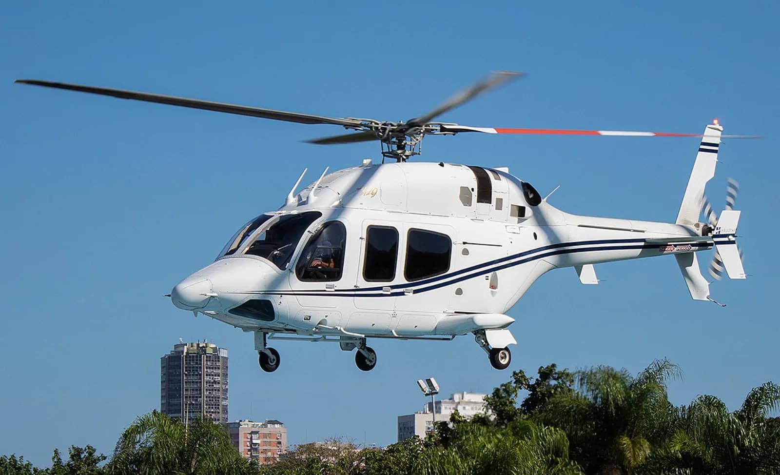 A white helicopter with blue stripes flying over a city with buildings and trees under a clear blue sky.
