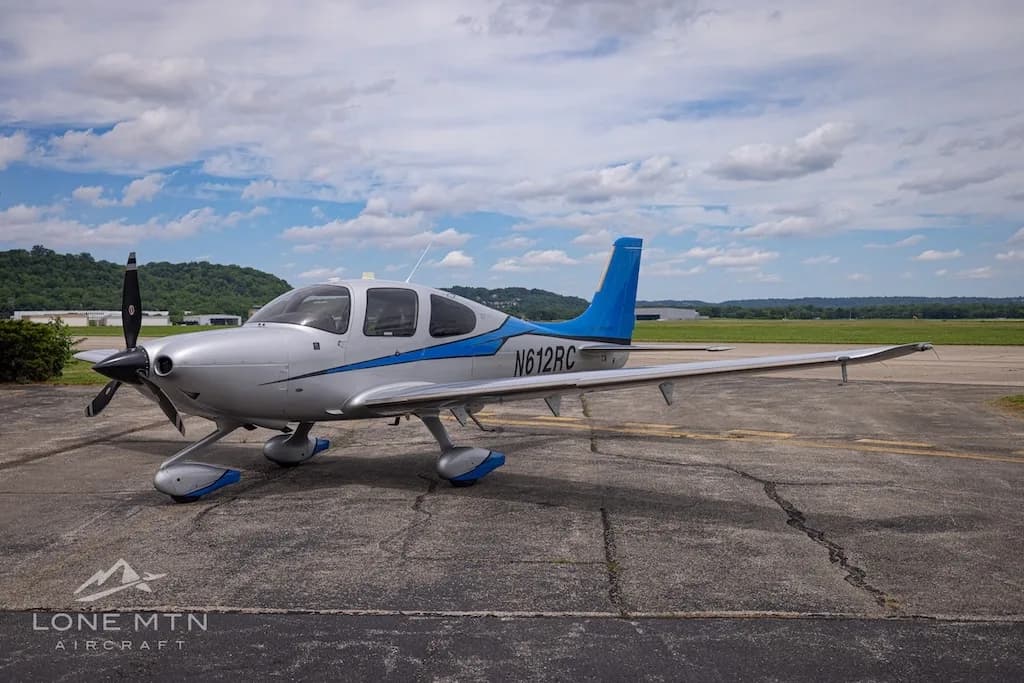 A silver and blue Cirrus SR22T Carbon aircraft, registration N612RC, parked on an airfield under a partly cloudy sky.