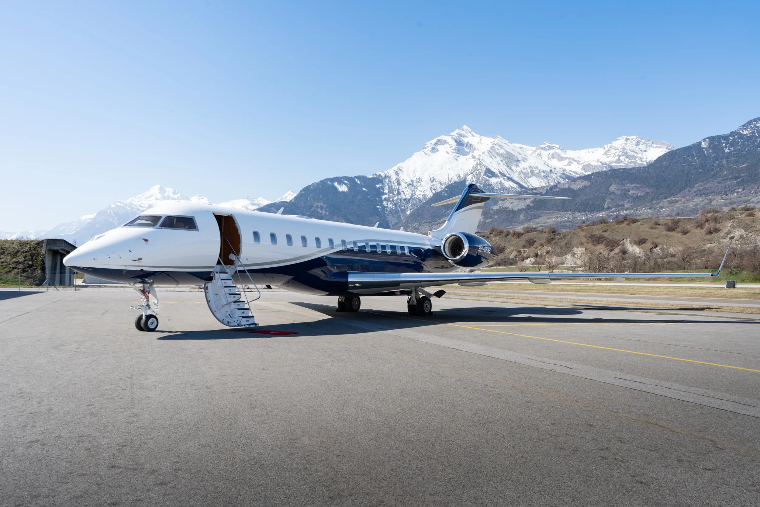 A white and dark blue private jet parked on an airfield with its door open, set against a backdrop of snow-capped mountains.