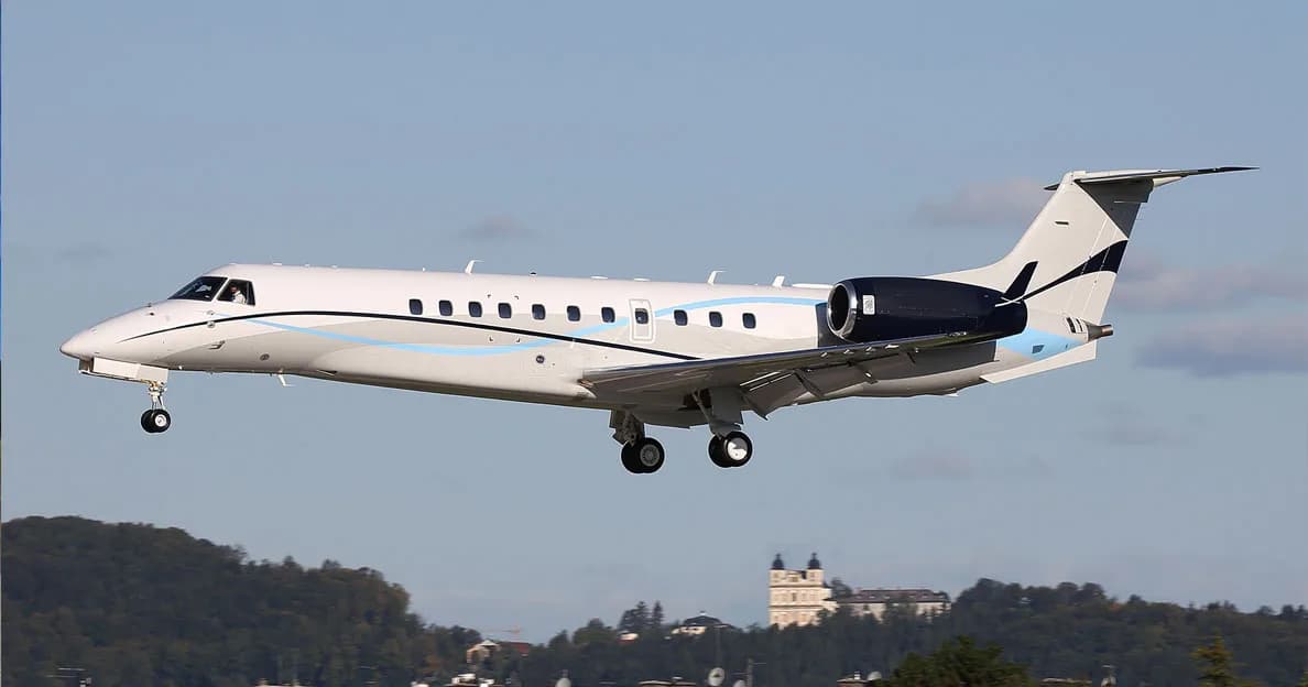 A white Embraer Legacy 600 series business jet with blue and black accents on its tail and engines is seen in flight, with landing gear extended, over a landscape with buildings.