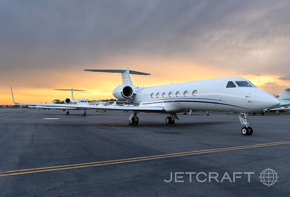 A white Gulfstream G550 business jet with blue stripes parked on a tarmac at sunset, with other aircraft in the background.