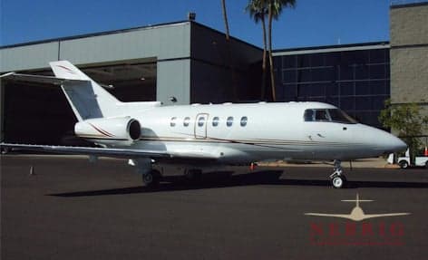 A white business jet with thin dark and light stripes is parked on an asphalt tarmac next to a hangar and building with palm trees.