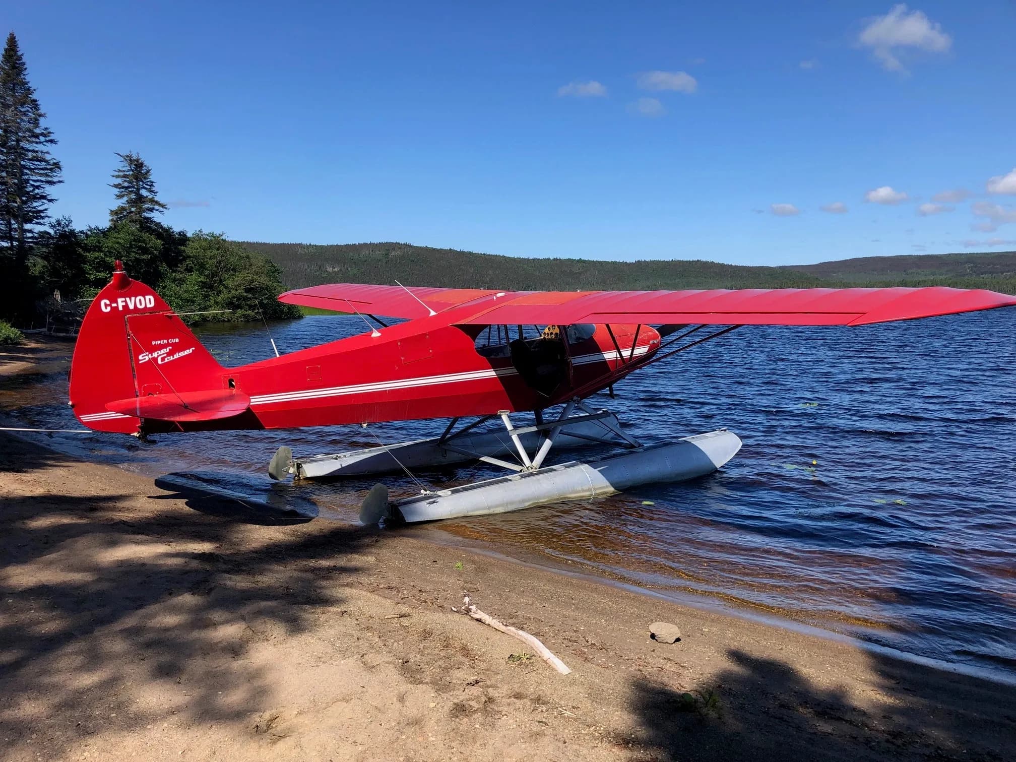 A red Piper Cub Super Cruiser seaplane (C-FVOD) with white stripes is partially beached on a sandy shore next to a lake, with trees and hills in the background under a blue sky.