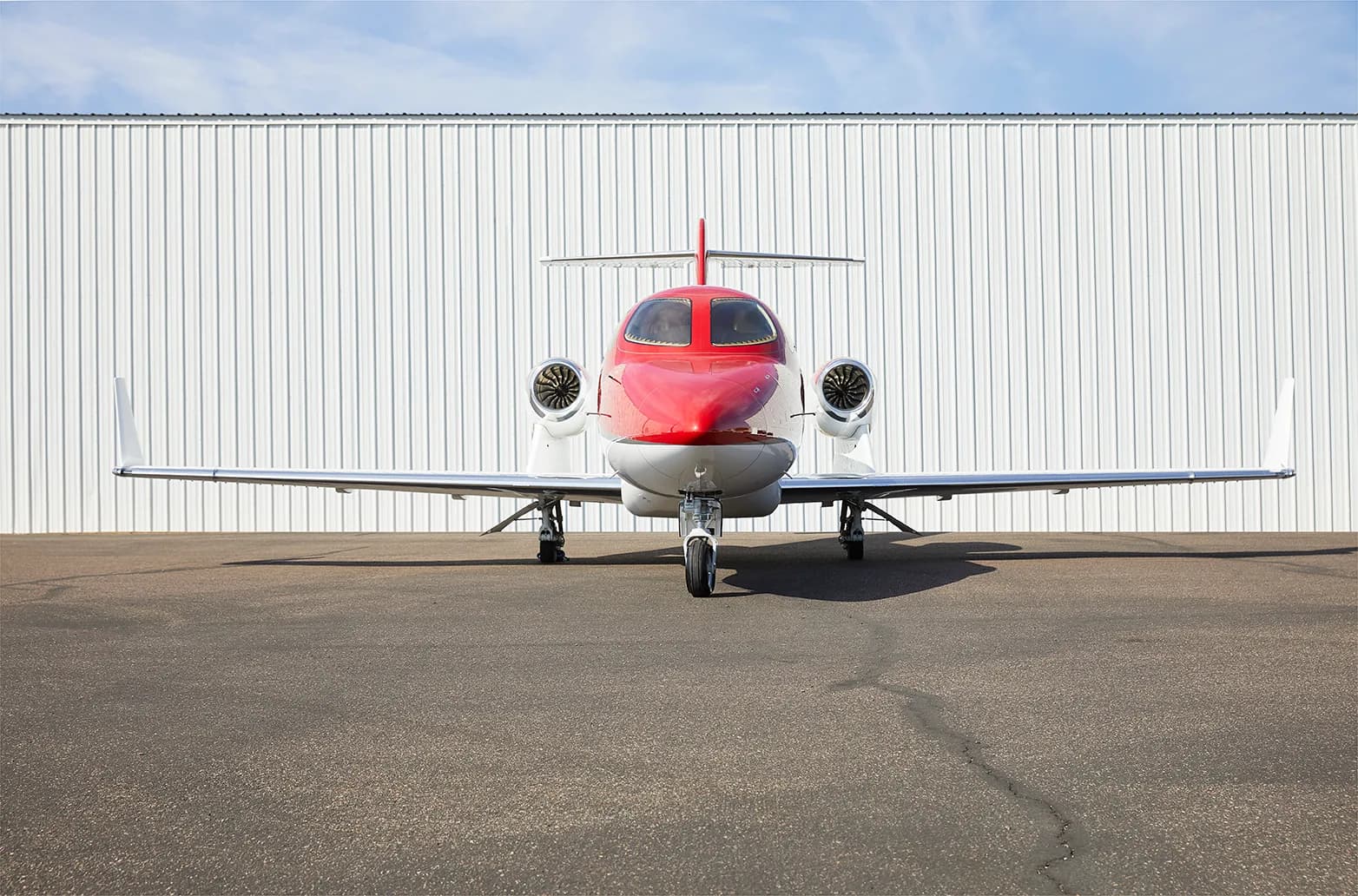A red and white HondaJet HA-420 aircraft is parked head-on on an asphalt tarmac in front of a white hangar.