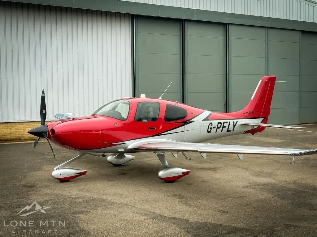 A red and white Cirrus SR22 aircraft (G-PFLY) parked on a tarmac in front of a hangar.