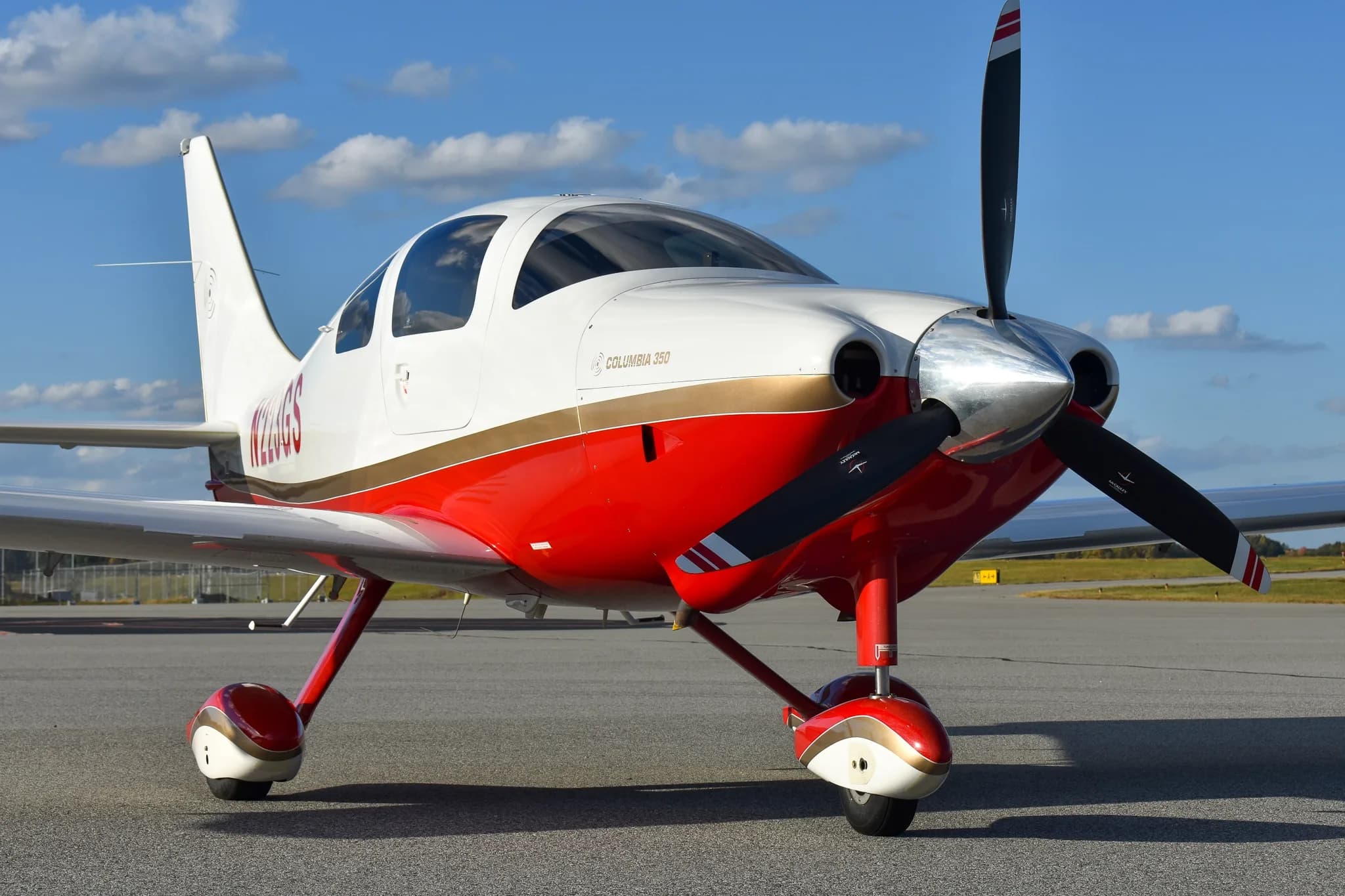Front-quarter view of a white and red Columbia 350 aircraft with a gold stripe, parked on an airfield under a blue sky.