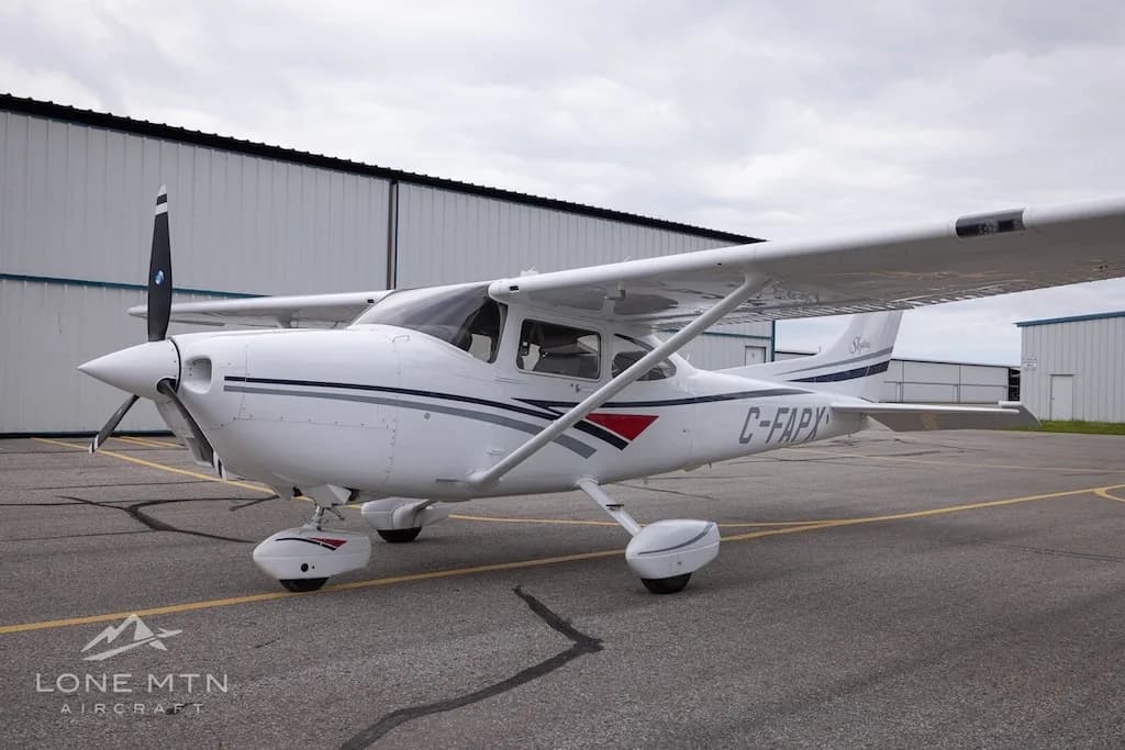 A white Cessna 182 Skylane with grey and blue stripes, registration C-FAPX, parked on an asphalt ramp in front of hangars.