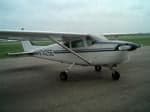 A white Cessna 172 with dark blue stripes is parked on an asphalt tarmac under a cloudy sky.
