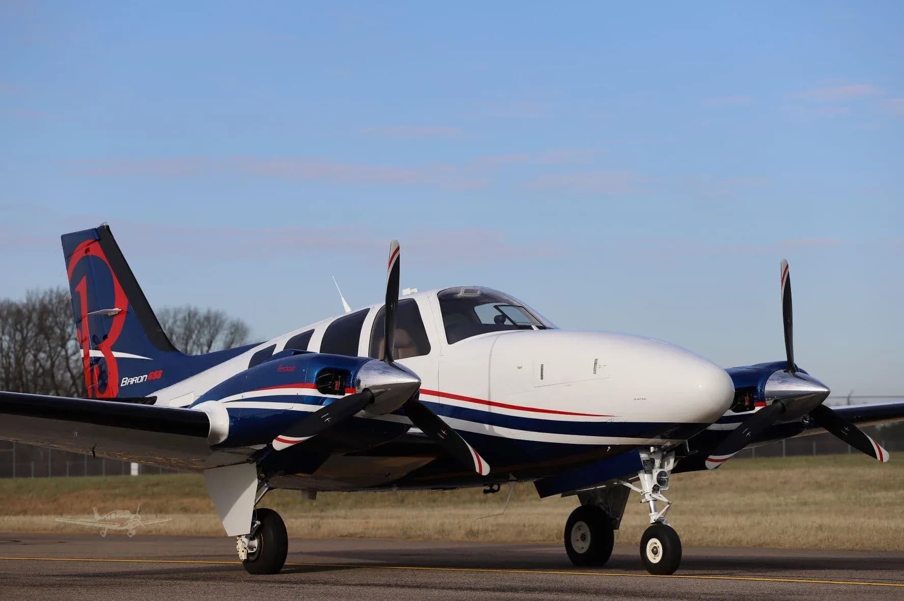 A white and blue Beechcraft Baron G58 with red accents on the tail, seen from the front-right on a tarmac under a partly cloudy sky.