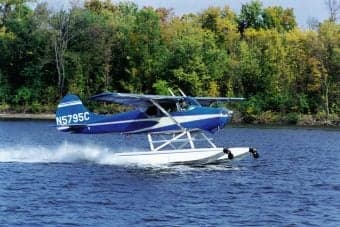 A blue and white seaplane, tail number N5795C, takes off from the water.