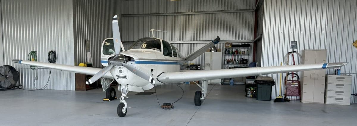 A white and blue Mooney M20K aircraft is parked inside a hangar with its doors open.