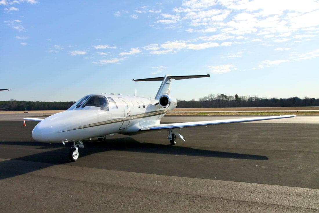 A Citation Jet sits on the tarmac in front of trees under a blue sky.