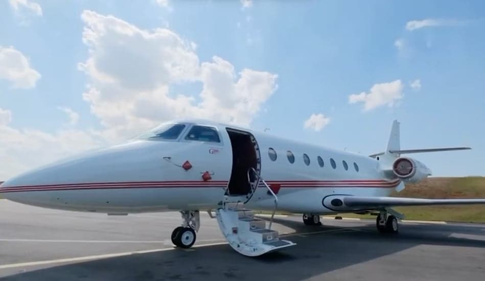 A white business jet with red stripes on the fuselage, parked on an tarmac with its main door open and stairs deployed.