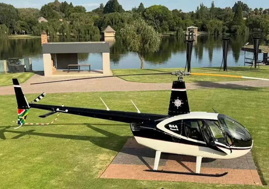 A black and white Robinson R44 Raven II helicopter with a South African flag on its tail boom is parked on a paved area next to a pond.