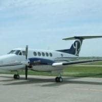 A white and blue twin-turboprop aircraft with a dark blue tail logo is parked on an airfield.