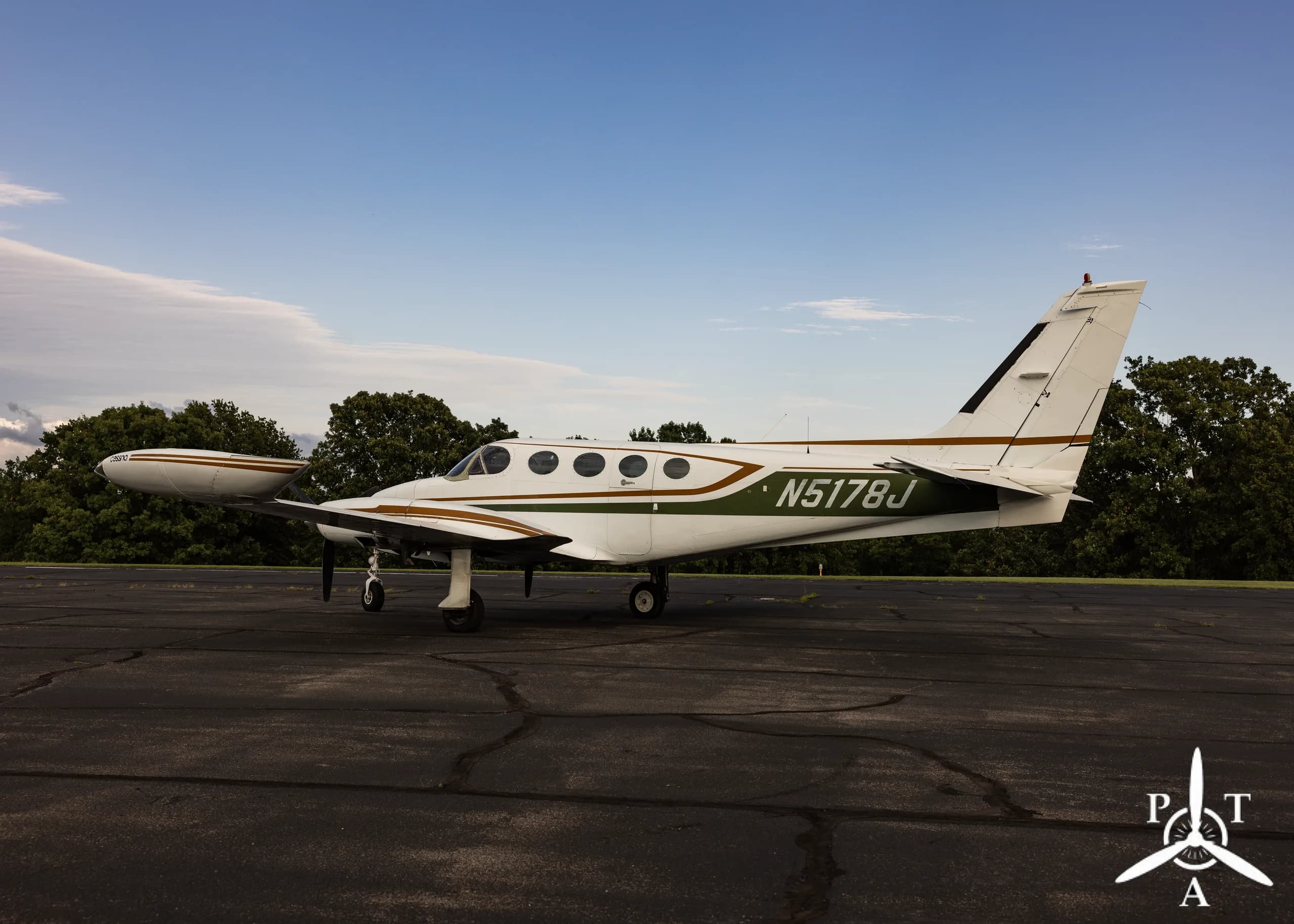 A white and green Cessna 340A twin-engine aircraft, N5178J, parked on an asphalt ramp with trees in the background under a blue sky.