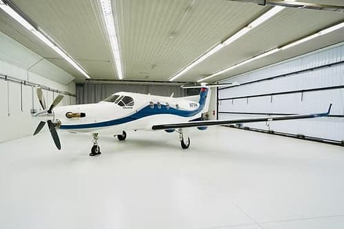 A Pilatus PC-12 aircraft with a white and blue paint scheme is parked inside a hangar.