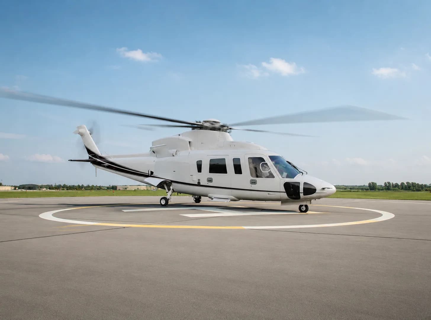 A white helicopter with black stripes sits on a helipad with its rotors spinning, under a blue sky.