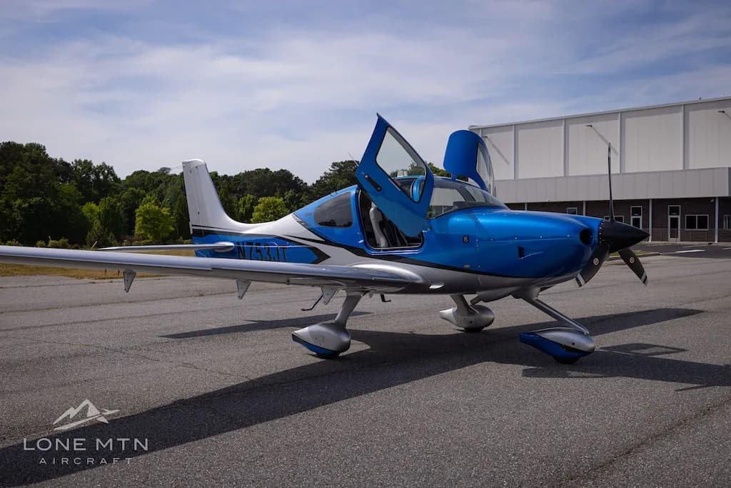 A blue and silver single-engine aircraft with open gull-wing doors is parked on an asphalt tarmac.