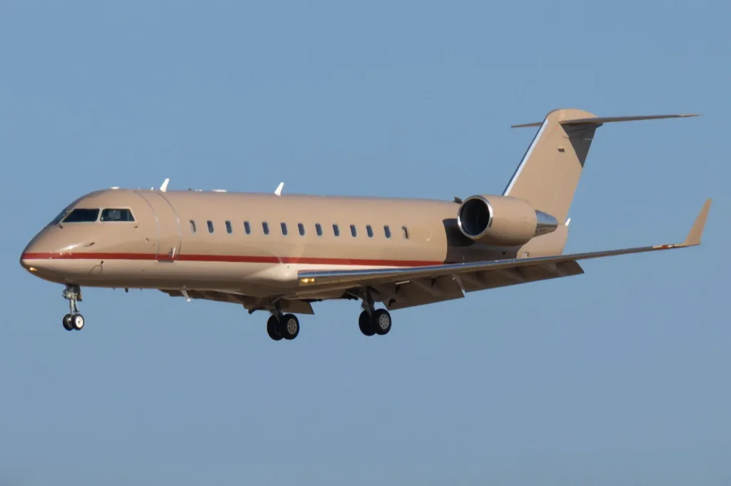 A beige private jet with a red stripe on its fuselage flies with landing gear extended against a clear blue sky.