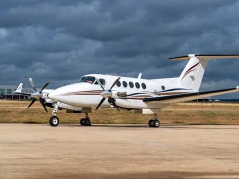A white twin-turboprop aircraft with brown and gold stripes on the fuselage and tail, parked on an asphalt tarmac under a cloudy sky.